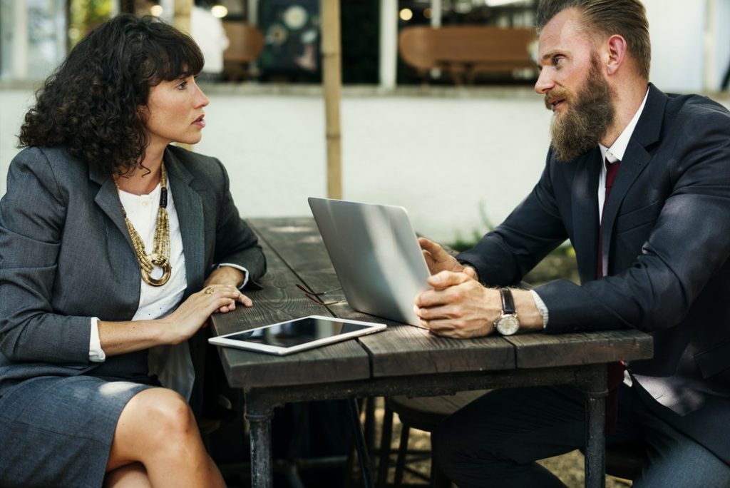 man and woman in business attire sit around a table with a laptop discussing something.