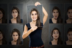 Is NLP is the ‘only way’ to deliver meaningful change? Or Is loyalty to the first change system you learnt a hindrance? Photograph of a young woman holding a smiling face over her face with photographs of her face with lots of other emotions in the background