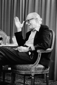 black and white photograph of an elderly but active looking white man wearing glasses, sitting in an armchair facing sideways and talking to someone out of sight.